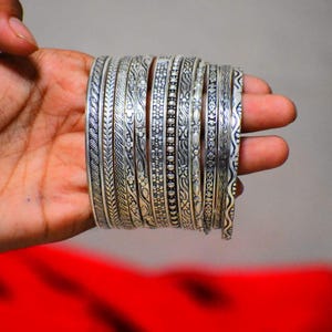 Op de afbeelding: Een set van tien zilverkleurige metalen armbanden met diverse reliëfpatronen, waaronder bloemmotieven, geometrische lijnen, gevlochten texturen en kraaldetails. Elke armband heeft een uniek ontwerp met geoxideerde accenten voor diepte.