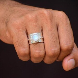 May include: A wide silver band ring featuring a central, circular, iridescent moonstone cabochon. The metal band has a smooth, polished finish and a distinct horizontal groove running along the center, encircling the finger with a solid structure.