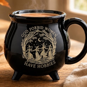 May include: A black ceramic cauldron-shaped mug with three small feet and a handle. The front features a beige graphic of four figures in pointed hats dancing under a crescent moon and stars, with the text "GOD FORBID WOMEN HAVE HOBBIES".