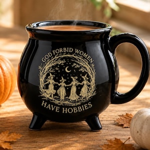 May include: A black ceramic cauldron-shaped mug with a handle and three feet. The front features a gold-toned illustration of four figures in hats dancing in a circle under a crescent moon, with the text "GOD FORBID WOMEN HAVE HOBBIES".
