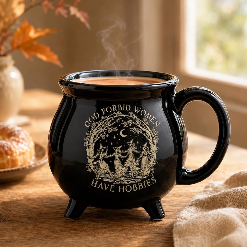 May include: A black ceramic cauldron-shaped mug with three small feet and a handle. The front features a beige graphic of four figures in pointed hats dancing under a crescent moon and stars, with the text "GOD FORBID WOMEN HAVE HOBBIES".
