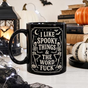 Op de afbeelding: Een zwarte keramische mok met een wit grafisch ontwerp van twee skeletten, vleermuizen, sterren en een maansikkel. De tekst luidt: I LIKE SPOOKY THINGS & THE WORD FUCK. Het ontwerp is omlijst door een decoratieve rand met kleine pompoenen.