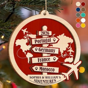May include: A round wooden Christmas ornament featuring a red background with a white world map, a central signpost with arrows pointing to Portugal, Germany, France, and Morocco, the year 2026, and the text Sophia & William's Adventures.
