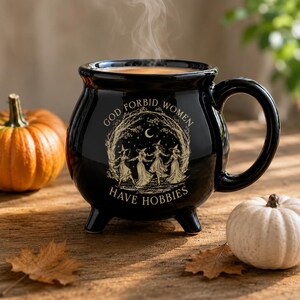 May include: A black ceramic cauldron-shaped mug with three small feet and a handle. The front features a gold-toned illustration of four figures in hats dancing under a crescent moon, with the text "GOD FORBID WOMEN HAVE HOBBIES" printed in a circular arrangement around the graphic.