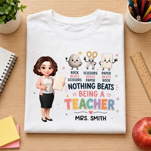 May include: A white t-shirt featuring a cartoon illustration of a teacher holding a book, alongside graphics of a rock, scissors, and paper. The text reads: Rock beats scissors, scissors beats paper, paper beats rock, nothing beats being a teacher, Mrs. Smith.