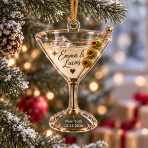 May include: A clear acrylic martini glass ornament with gold trim, featuring three green olives with red centres on a pick. The glass is engraved with the text Emma & Lucas, a heart, and the text New York 12.14.2026, suspended by a gold cord.