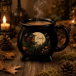 May include: A black ceramic cauldron-shaped mug with three small feet and a handle. The front features a graphic of a witch on a broomstick against a full moon, surrounded by forest elements and the name Emily written in cursive script.