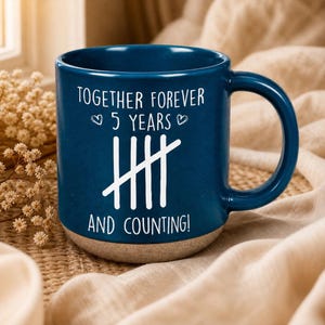 May include: A dark blue ceramic mug with a speckled light brown base. The front features white text that reads "TOGETHER FOREVER 5 YEARS AND COUNTING!" with two small heart icons and a tally mark graphic representing the number five in the center.