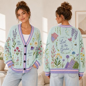 May include: A light blue V-neck cardigan with purple striped trim, featuring embroidered wildflowers and the text Softly Bloom Meadow Notes on the front, and Lost in the Garden with floral and insect embroidery on the back.