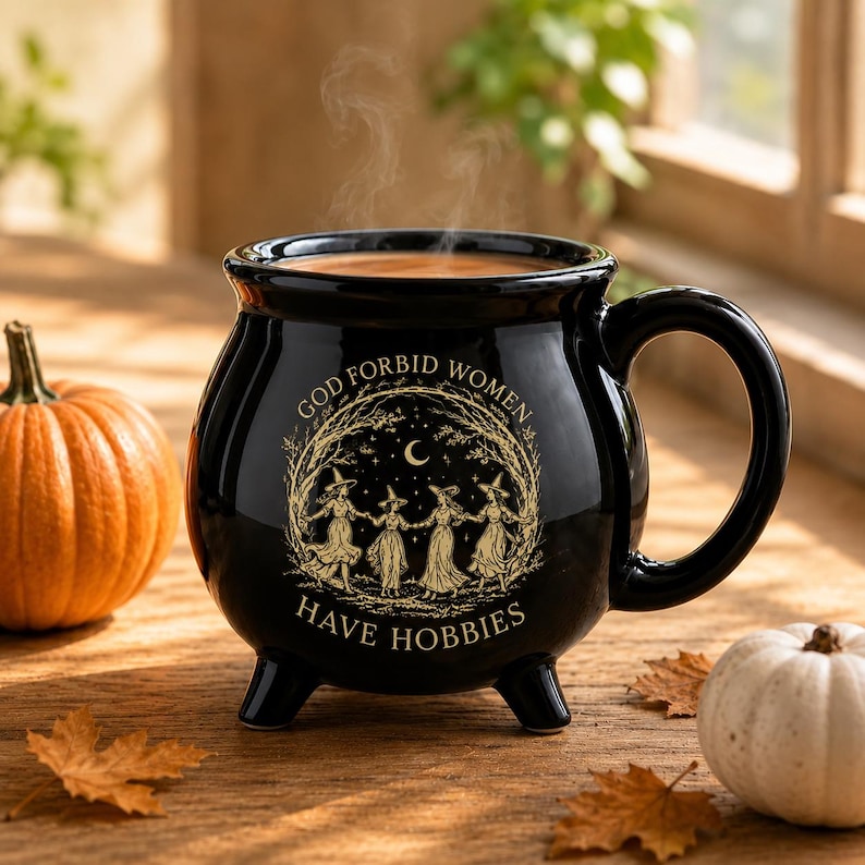 May include: A black ceramic cauldron-shaped mug with a handle and three feet. The front features a gold-toned illustration of four figures in hats dancing in a circle under a crescent moon, with the text "GOD FORBID WOMEN HAVE HOBBIES".