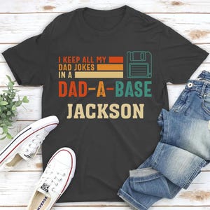 Könnte beinhalten: Ein dunkelgraues Kurzarm-T-Shirt mit einer Grafik einer Diskette und dem Text I KEEP ALL MY DAD JOKES IN A DAD-A-BASE JACKSON in Orange, Gelb und Türkis. Das Design enthält horizontale Balken und eine Schriftart im Retro-Stil.