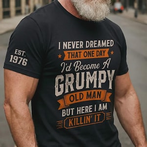 Könnte beinhalten: Ein schwarzes Kurzarm-T-Shirt mit orangefarbenem und weißem Text, der lautet I NEVER DREAMED THAT ONE DAY I'D BECOME A GRUMPY OLD MAN BUT HERE I AM KILLIN' IT. Auf dem rechten Ärmel steht der Text EST. 1976 in weißer Schrift.