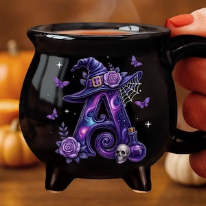 Pode incluir: Uma caneca preta em forma de caldeirão com uma letra A grande e roxa decorada com um chapéu de bruxa, rosas, borboletas, uma teia de aranha, um pequeno crânio e um frasco de poção. A letra tem um padrão de redemoinho galáctico.