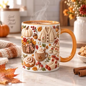 May include: A white ceramic mug with a glossy orange handle and rim, featuring raised 3D-style decorations including a pumpkin, a woven basket of autumn leaves, a knit cardigan, mittens, socks, a cinnamon roll, and a small mug of hot cocoa.
