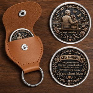 May include: A tan leather keychain pouch with a silver snap button and metal ring, alongside two circular metal medallions. One medallion depicts a grandmother and granddaughter on a bench with the text TO MY GRANDDAUGHTER, GRANDMA, SOPHIA, and I Love You. The other features the text LIFE WILL BLOOM, KEEP GROWING, and a sentimental message signed LOVE, GRANDMA.