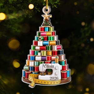 May include: A Christmas tree-shaped ornament constructed from stacked colorful thread spools. At the base sits a white sewing machine featuring the name Olivia and the year 2026, wrapped in a yellow measuring tape. A gold star tops the tree.