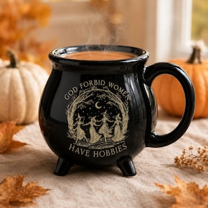 May include: A black ceramic cauldron-shaped mug with three small feet and a handle. The front features a gold-toned illustration of four figures in pointed hats dancing in a circle under a crescent moon, with the text GOD FORBID WOMEN HAVE HOBBIES.