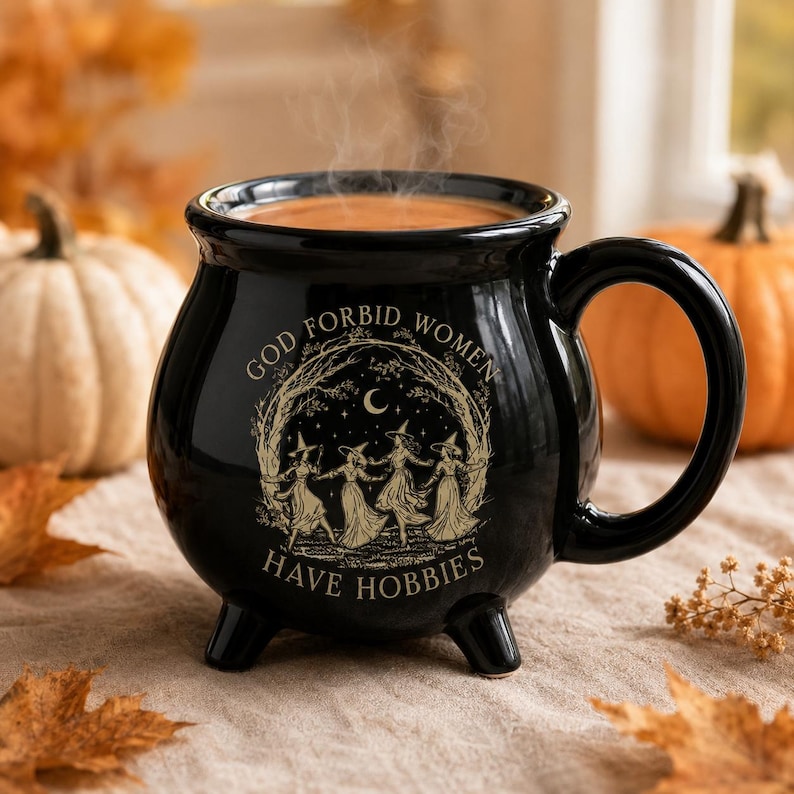 May include: A black ceramic cauldron-shaped mug with three small feet and a handle. The front features a gold-toned illustration of four figures in pointed hats dancing in a circle under a crescent moon, with the text GOD FORBID WOMEN HAVE HOBBIES.