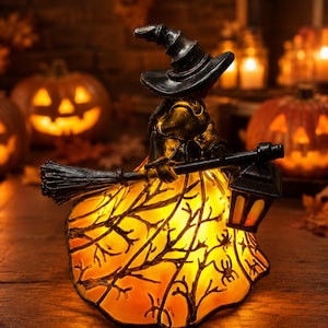 May include: A decorative witch figurine featuring a black pointed hat and a flowing dress illuminated from within. The dress displays a dark silhouette pattern of bare tree branches and spiders. The figure holds a broom and a black lantern.