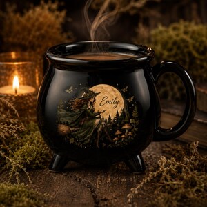 May include: A black ceramic cauldron-shaped mug with a handle and three small feet. The front features a graphic of a person in a hat riding a broomstick in front of a large full moon, with the name Emily printed in script, mushrooms, and butterflies.