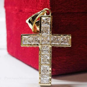 Puede incluir: Un colgante de cruz de oro con una serie de piedras transparentes de corte cuadrado dispuestas en cuadrícula. El colgante incluye una anilla de oro pulido en la parte superior para sujetarlo a una cadena, con un acabado metálico brillante.