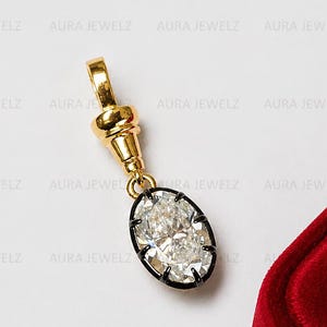 May include: A gold pendant featuring an oval-shaped faceted clear stone set in a dark metal frame. The pendant is attached to a gold bail with a cylindrical connector. The piece is positioned next to a red velvet-textured jewelry box corner.