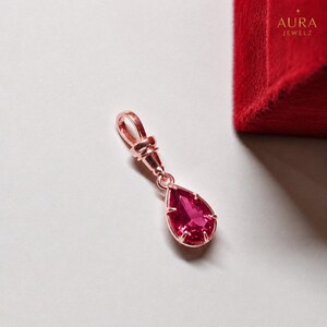 May include: A rose gold pendant featuring a pear-shaped, faceted deep pink gemstone held by four prongs. The pendant includes a polished rose gold bail at the top for attachment. The text AURA JEWELZ is visible in the upper right corner.