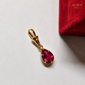 May include: A gold pendant featuring a pear-shaped, faceted deep pink gemstone held by four prongs. The pendant includes a polished gold bail at the top for attachment. The piece is positioned near a red velvet surface with the text AURA JEWELZ.