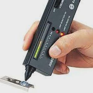 May include: A black handheld diamond tester device with a pointed probe tip, a vertical LED indicator scale, and printed text including "DIAMOND SELECTOR II". The device is positioned over a small, clear, faceted gemstone set in a metal base.