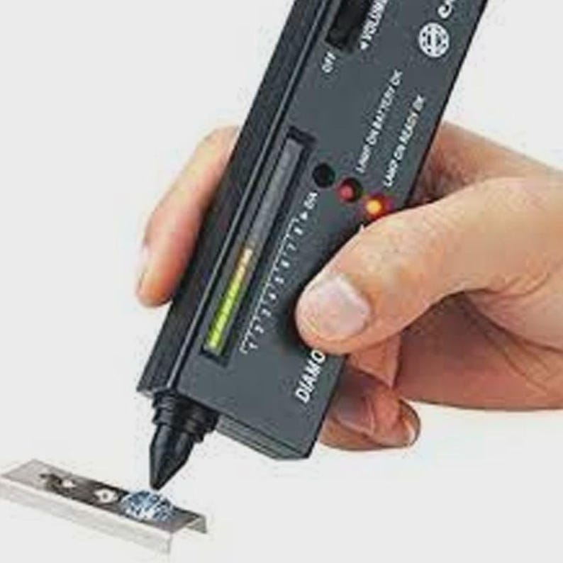 May include: A black handheld diamond tester device with a pointed probe tip, a vertical LED indicator scale, and printed text including "DIAMOND SELECTOR II". The device is positioned over a small, clear, faceted gemstone set in a metal base.