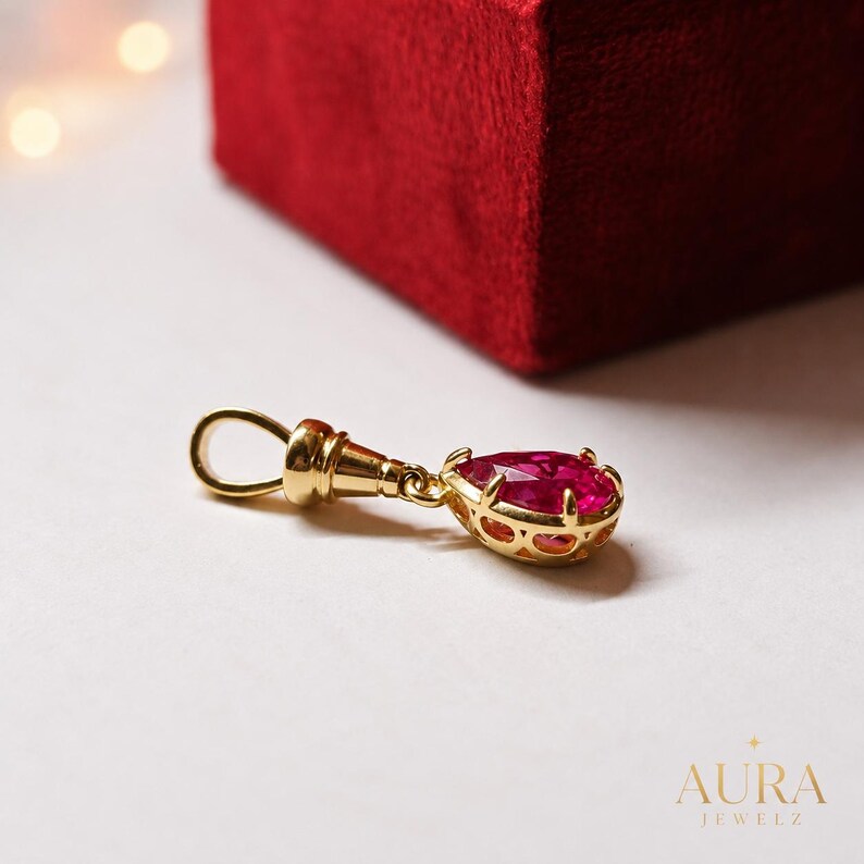 May include: A gold pendant featuring a pear-shaped, faceted deep pink gemstone set in a decorative gold cage. The pendant includes a polished gold bail at the top for attachment to a chain, showcasing intricate metalwork around the stone setting.