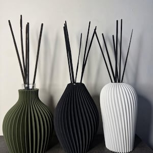 Pode incluir: Três difusores de varetas com corpos canelados e bulbosos em verde-oliva, preto fosco e branco. Cada recipiente contém um grupo de varetas pretas finas que se estendem para cima a partir do gargalo estreito, criando um conjunto uniforme.