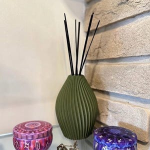 Pode incluir: Um vaso difusor de varetas verde-oliva fosco com varetas pretas, uma pequena figura de bicicleta metálica e duas latas redondas de metal com padrões do zodíaco em roxo e rosa com símbolos astrológicos brancos impressos nas tampas.