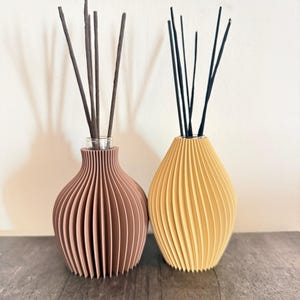 Puede incluir: Dos difusores de varillas con recipientes acanalados en forma de jarrón. Un recipiente es de color marrón terracota mate y el otro es amarillo mate. Cada vasija contiene un grupo de varillas finas de color marrón oscuro hacia arriba.