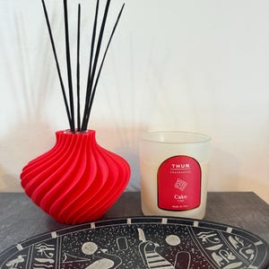 Può includere: Un diffusore a bastoncini rosso brillante a forma di spirale con bastoncini neri, una candela in vetro satinato con etichetta rossa recante la scritta THUN FRAGRANCES Cake TIME MADE IN ITALY e un piatto decorativo nero con motivi geroglifici.