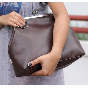 May include: A dark brown clutch bag with a smooth, matte finish and a silver-toned metal kiss-lock frame closure. The bag features a small rectangular brand tag on the side and a structured, wide base design for holding personal items securely.