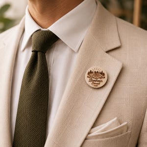 Peut inclure: Gros plan d'une personne portant un costume beige, une chemise blanche et une cravate vert olive. Une épingle en bois avec les mots "Bonheur Éternel - 2026" est épinglée sur le revers. L'épingle présente un motif de montagne et d'arbres.
