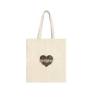 May include: Cream-colored tote bag with a heart-shaped patch in the center. The patch is made of various plaid fabrics in shades of brown, green, and yellow. The word "OUTSIDER" is printed in white across the heart.