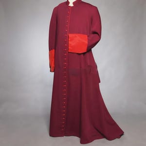 May include: A floor-length burgundy cassock featuring a high mandarin collar and a long vertical row of matching fabric-covered buttons down the center front. The garment includes contrasting bright red cuffs on the long sleeves.