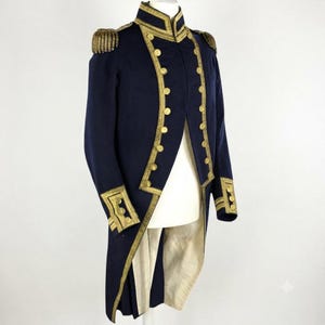 Può includere: Una giacca a coda in stile militare blu navy scuro con ricami metallici dorati lungo il colletto, l'apertura frontale e i polsini. Il capo presenta bottoni color oro, spalline strutturate con frange e una fodera di colore chiaro.