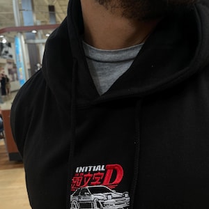 Può includere: Felpa con cappuccio nera con logo "INITIAL D" ricamato, raffigurante un'auto bianca con dettagli rossi. La felpa ha un cappuccio con coulisse e si intravede una maglietta grigia. Primo piano.
