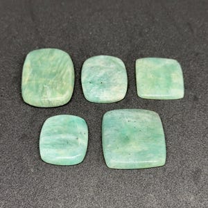 May include: Five polished, rectangular amazonite gemstones with rounded corners. Each stone displays a unique pattern of pale mint green and white marbling with subtle dark inclusions, showcasing a smooth, reflective surface and flat, cabochon-like shape.