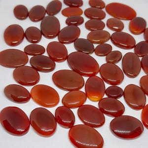May include: A collection of numerous smooth, polished, oval-shaped carnelian gemstones in various shades of deep orange, reddish-brown, and burnt sienna. Each stone features a glossy, reflective surface with subtle variations in colour and translucency.