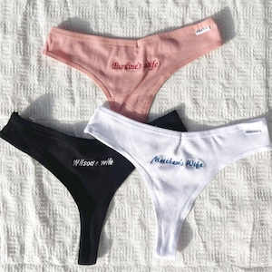 May include: Three ribbed thong panties in pink, black, and white. The pink pair features red embroidery reading Burdine's wife, the black pair has white embroidery reading Wilson's wife, and the white pair has blue embroidery reading Matthew's Wife.