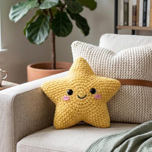 May include: A yellow star-shaped crochet cushion with a smiling face, black eyes, and pink cheeks sits on a beige sofa. A cream-coloured cushion with a brown leather strap and a green blanket are also on the sofa. A coffee cup is on a side table.