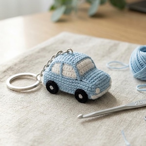 May include: A light blue and white crocheted car keychain with black wheels and a silver metal ring. A silver crochet hook with 2.0 mm text is visible nearby, along with a small ball of light blue yarn and a metal stitch marker on the fabric.