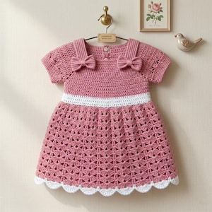 Può includere: Un abito all'uncinetto a maniche corte in rosa polvere con fascia in vita bianca e orlo smerlato bianco. Il corpetto presenta due piccoli fiocchi sulle spalle e una chiusura con bottoni sul retro. La gonna ha un motivo a conchiglia testurizzato.