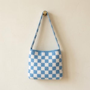 Könnte beinhalten: Eine handgefertigte, blau-weiß karierte Häkeltasche. Die Tasche hat eine quadratische Form und einen passenden Riemen. Das Muster ist ein sich wiederholendes Schachbrettmuster. Die Tasche hängt an einer Wand.