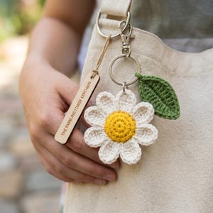 May include: A handmade daisy keychain with a yellow center and white petals, paired with a green crocheted leaf. The keychain includes a wooden tag that reads "THE YARNLLNG | DAISY KEYCHAIN". The keychain is attached to a silver metal ring and clasp.