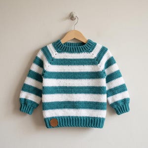 Peut inclure: Un pull crocheté à rayures blanches et bleu sarcelle, présenté sur un cintre en bois. Le pull présente des rayures horizontales et une encolure ronde et côtelée. Un petit patch rond en cuir marron est cousu sur le côté inférieur gauche du pull.